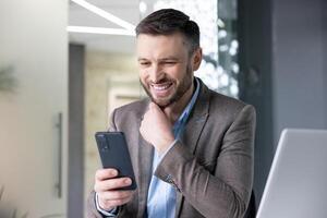 alegre sonriente satisfecho empresario mecanografía texto mensaje en teléfono, hombre en negocio traje hojeada social medios de comunicación y utilizando aplicación en teléfono inteligente, sentado dentro oficina con ordenador portátil a lugar de trabajo. foto