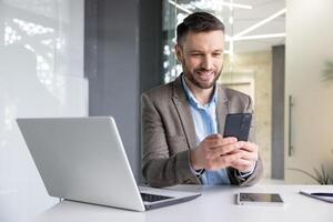 alegre sonriente satisfecho empresario mecanografía texto mensaje en teléfono, hombre en negocio traje hojeada social medios de comunicación y utilizando aplicación en teléfono inteligente, sentado dentro oficina con ordenador portátil a lugar de trabajo. foto