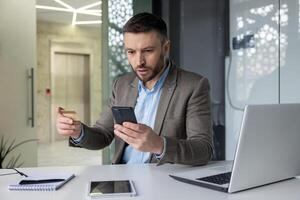 Businessman discovering online fraud while looking at his smartphone in an office setting. The image captures the concept of financial fraud, caution, and security. photo