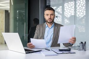 Surprised businessman holding documents while working at his desk in a modern office setting. Expression of shock and concern. photo