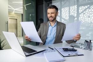 retrato exitoso financiero contador a lugar de trabajo dentro oficina, hombre aspecto, cheques informes, sostiene contrato documentos en manos, empresario en negocio traje sonriente, satisfecho logro resultados. foto