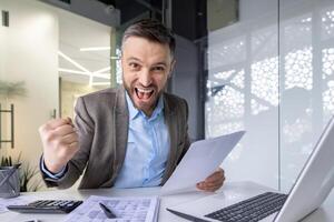 A triumphant businessman in a suit celebrates success with a fist pump at his office workspace, surrounded by documents. photo