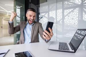 empresario celebrando victoria triunfo exitoso logro resultados a lugar de trabajo dentro oficina, jefe en negocio traje participación teléfono, utilizando teléfono inteligente aplicación, recibido victoria notificación. foto