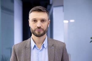 Confident businessman in office attire standing indoors with a neutral expression and modern office background. photo