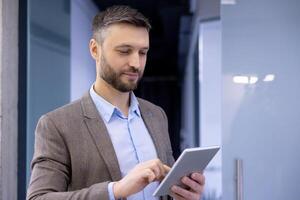 Professional man in smart casual outfit smiling while using a tablet in a modern office setting, depicting work and technology. photo