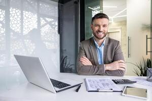 Portrait of successful financier accountant at workplace inside office, senior experienced businessman smiling looking at camera with crossed arms, working with papers sitting at table with laptop. photo