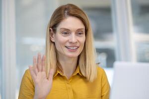 Attractive blonde woman in a yellow blouse smiling and waving at the camera during a call, sitting in a bright office space. photo