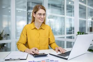 A professional young woman with glasses works on her laptop while using a calculator in a bright, contemporary office environment. photo