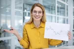 A cheerful businesswoman wearing glasses holding a pen and explaining financial charts in a bright, modern office setting. photo