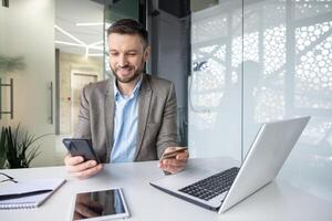 joven empresario haciendo en línea pago utilizando teléfono inteligente y crédito tarjeta a un moderno oficina escritorio con un computadora portátil, tableta, y bloc. foto