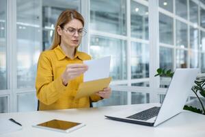 A focused businesswoman in a modern office reading documents from a yellow envelope, displaying a confused and bothered expression. photo