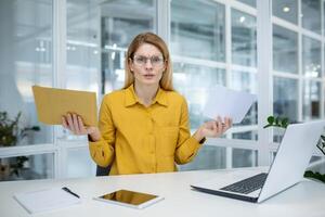 Confused businesswoman wearing yellow shirt holding postal envelopes in a modern office setting, expressing frustration or confusion. photo