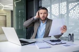 Shocked businessman holding a document while sitting at his desk in an office. He appears surprised and worried, surrounded by paperwork and a laptop. photo