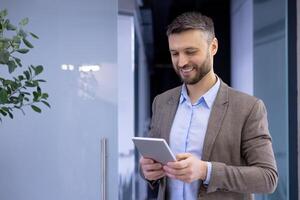 Mature experienced businessman inside office smiling contentedly, senior man holding tablet computer in hands, boss in business suit using analytical financial application. photo