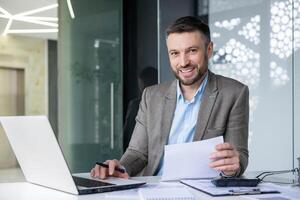Smiling businessman at his desk working on laptop and checking documents in a contemporary office setting. photo