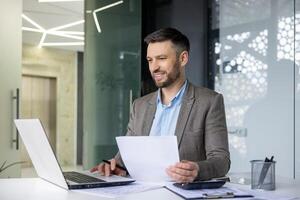 Confident businessman working in a modern office, holding documents and using a laptop. Professional workspace with contemporary design. photo