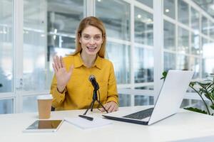 sonriente mujer en un amarillo camisa hospedaje un podcast desde su oficina escritorio, presentando un ordenador portátil y un micrófono. profesional preparar para en línea contenido creación. foto