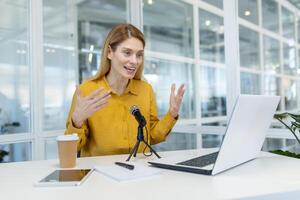 Engaged female podcast host interacts with audience using a mic and laptop in a bright office space. She appears enthusiastic and professional. photo