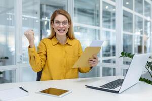 Joyful woman receiving positive news through a letter in the office, celebrating with a fist pump. Modern office environment. photo