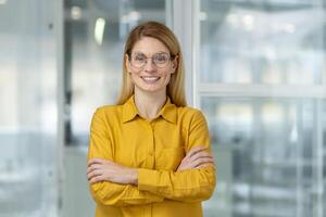 Portrait of a cheerful, confident businesswoman wearing a bright yellow blouse and glasses, standing with arms crossed in a modern office setting. photo