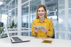 Joyful businesswoman counting money while working at office desk with a laptop and tablet. Concept of success, financial gain, and workplace accomplishment. photo