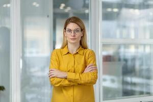 Confident businesswoman with glasses standing in modern office, arms crossed, looking at camera. Professional and determined appearance in contemporary workspace. photo