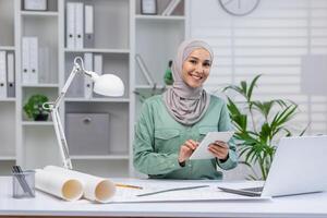 Confident female architect with hijab using a tablet in a modern office, surrounded by design papers and a laptop. photo