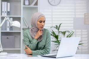 Woman wearing hijab focused while working on a laptop in a well-lit, modern office setting. Professional and thoughtful workspace environment. photo