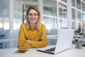 Smiling woman in a modern office setting, sitting at a desk with a laptop, wearing glasses and a yellow blouse with confidence. photo