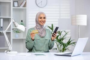 Confident woman wearing hijab using credit card and smartphone for online shopping at home office setup, modern business lifestyle concept photo