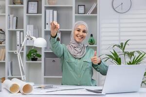 confidente hembra corredor de bienes raíces vistiendo un hijab sostiene casa llaves y da un pulgares arriba mientras trabajando en un moderno oficina. concepto de éxito, real bienes, y profesional logro. foto