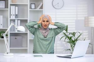 Stressed woman in hijab holding head in frustration while working in modern office. Concept of workplace anxiety, stress, and dealing with professional challenges. photo