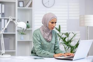 Frustrated woman in a hijab sitting at a desk while working on a laptop in a modern office setting. photo