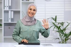 Confident woman wearing hijab waves while working from home office, engaging in virtual business meeting or online communication. photo