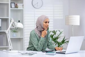 A focused woman in hijab working on a laptop in a modern office setting, demonstrating concentration and professionalism. photo