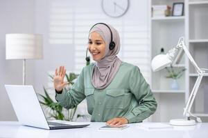 Smiling woman wearing a hijab using a laptop for a call while working from home in a modern office setup. photo