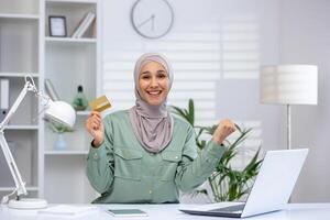 Smiling woman wearing hijab holding a credit card while working on a laptop at a modern home office. Successful online shopping and digital transactions concept. photo