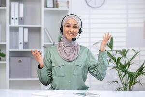 Smiling customer support representative wearing hijab and headset, working in a modern office setting with bookshelves and plants. photo