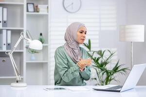 A concerned woman in hijab holding her wrist due to pain while working on a laptop in a bright, modern office setting. photo