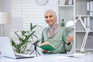 Confident woman in hijab hosting a podcast at her home desk. Holding a book in front of a microphone while smiling towards the camera. photo