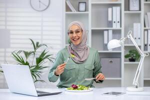 A joyful Muslim woman wearing a hijab eats a fresh salad at her desk, taking a break from work with her laptop and lamp beside. photo