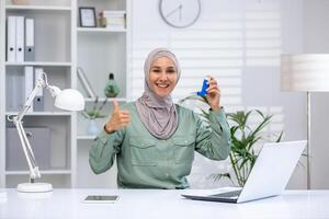 Confident woman in hijab working from home office, showing asthma inhaler and giving thumbs up gesture, concept of health and remote work photo
