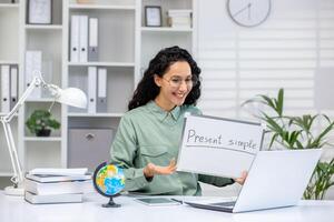 Smiling woman giving an online lesson using a whiteboard in a home office setting. Educational materials and a laptop are visible. photo