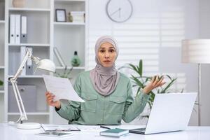 frustrado mujer de negocios vistiendo hijab participación documentos, asistiendo un en línea reunión en un moderno oficina. concepto de trabajo estrés, confusión, y negocio retos foto