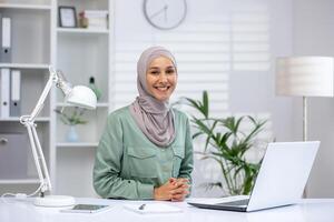 confidente mujer de negocios vistiendo hijab sentado a escritorio con ordenador portátil en brillante y moderno oficina ambiente. concepto de profesionalismo, diversidad, y éxito. foto