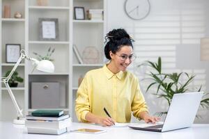 A smiling professional Hispanic woman engages in her work on a laptop at a well-organized home office. She exudes confidence and job satisfaction, symbolizing remote work opportunities. photo