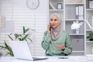 Confident Muslim businesswoman in a hijab speaking on mobile phone, expressing ideas in a modern office setting. photo