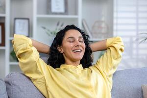 Close up of successful smiling and satisfied woman with hands behind head with closed eyes smiling and happy sitting on sofa, in living room at home in house inside. photo
