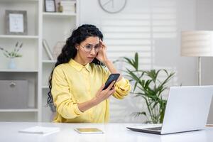 Perplexed caucasian female with long black curly hair holding personal mobile phone and sitting in front of open laptop on white desk. Wall clock in office or study room setting. photo