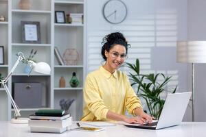 Portrait of happy successful woman at work in home office, hispanic woman smiling and looking at camera sitting at table in living room, working with laptop remotely, studying online. photo